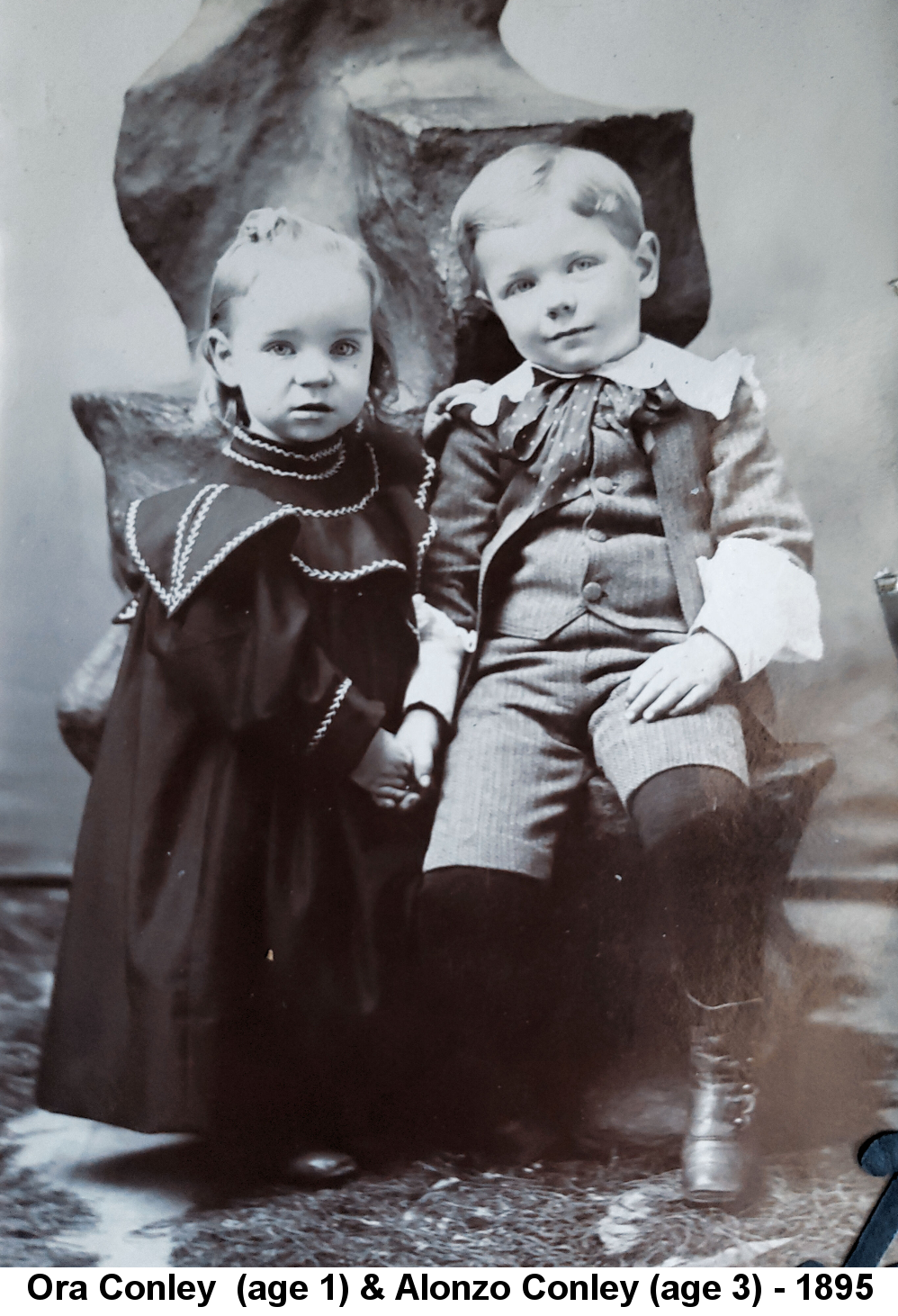 IMAGE/PHOTO: Ora Conley (age 1) & Alonzo Conley (age 3) - 1895: Black and white studio photo of two small children; on the left a little girl with blonde hair stands in front of a large rock on a ledge of which sits a little boy, also with blond hair, and they are holding hands.